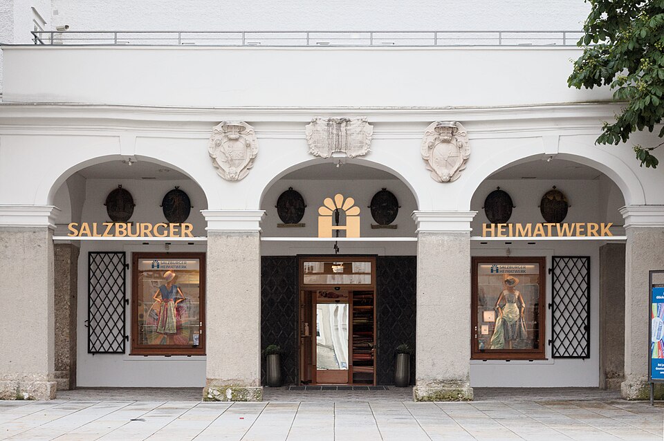 一棟白色歷史建築的拱廊立面，門面上方橫向排列著 SALZBURGER HEIMATWERK 字樣，兩側為鑲鉛玻璃的展示窗。
