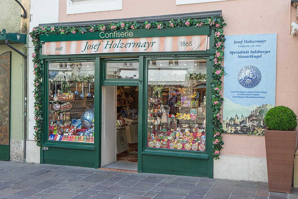 一間綠色門面的老糖果店，招牌寫著 Confiserie Josef Holzermayr 與 1888，櫥窗內陳列大量包裝商品。