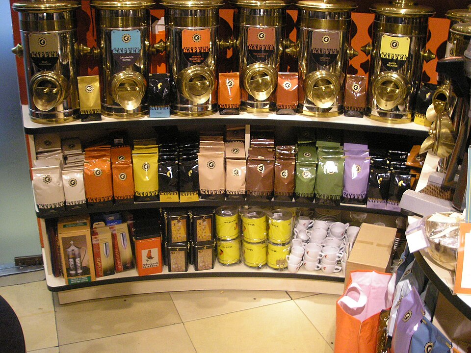 店內弧形貨架上陳列成排的 Julius Meinl 咖啡罐與包裝，罐身有橘、綠、紫等不同顏色標示不同品項。