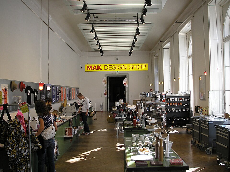 狹長明亮的博物館商店空間，黃色招牌寫著 MAK DESIGN SHOP，兩側展示櫃陳列書籍與設計商品。