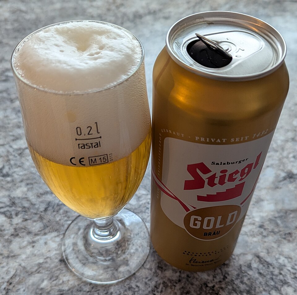 大理石桌面上一杯滿是泡沫的金黃色啤酒與一罐 Stiegl Gold 啤酒罐並排。