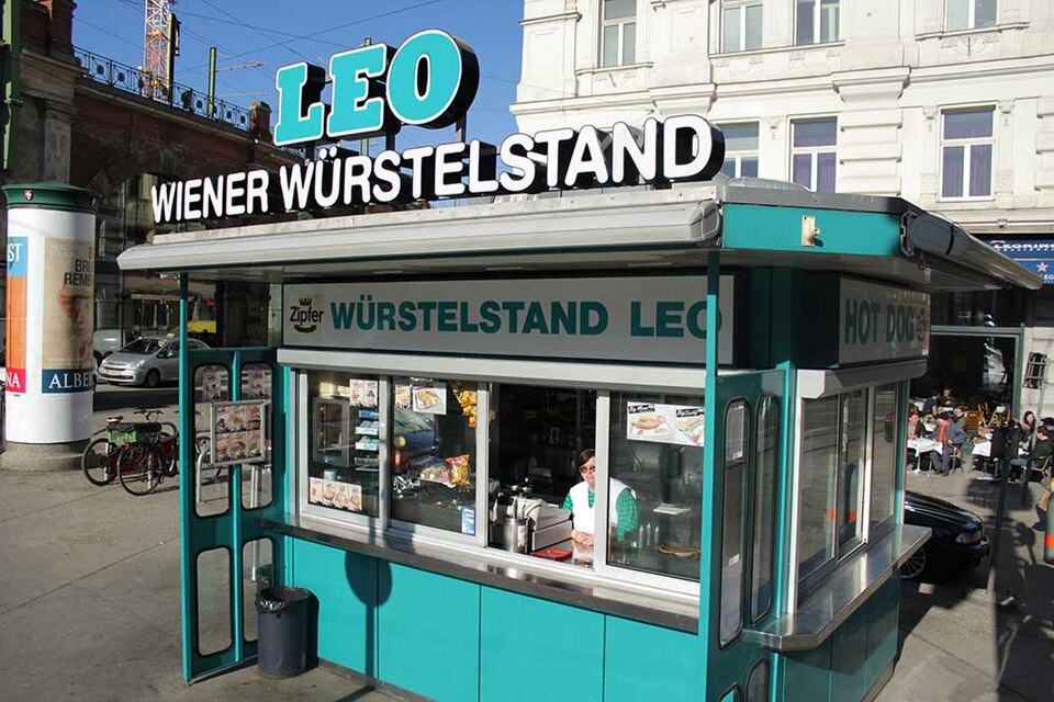 街邊一座青綠色的方形小攤，招牌上寫著 LEO WIENER WÜRSTELSTAND，攤內有人正在製作食物。